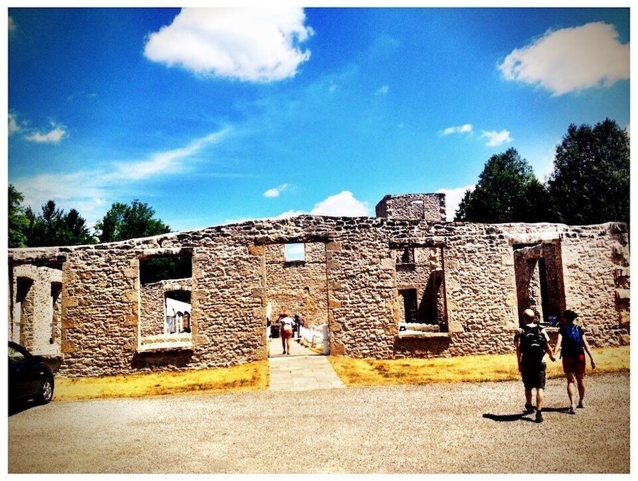 Ein altes Steinhaus mit Rundbögen und Fenstern, umgeben von Bäumen und wolkenlosem, blauem Himmel.