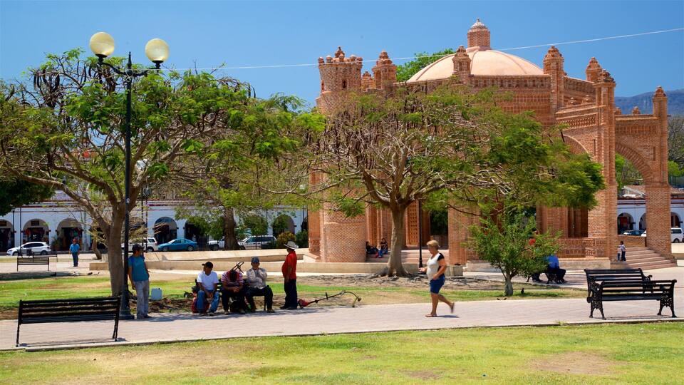 A park with a large brick building in the background, benches, and people sitting and walking.