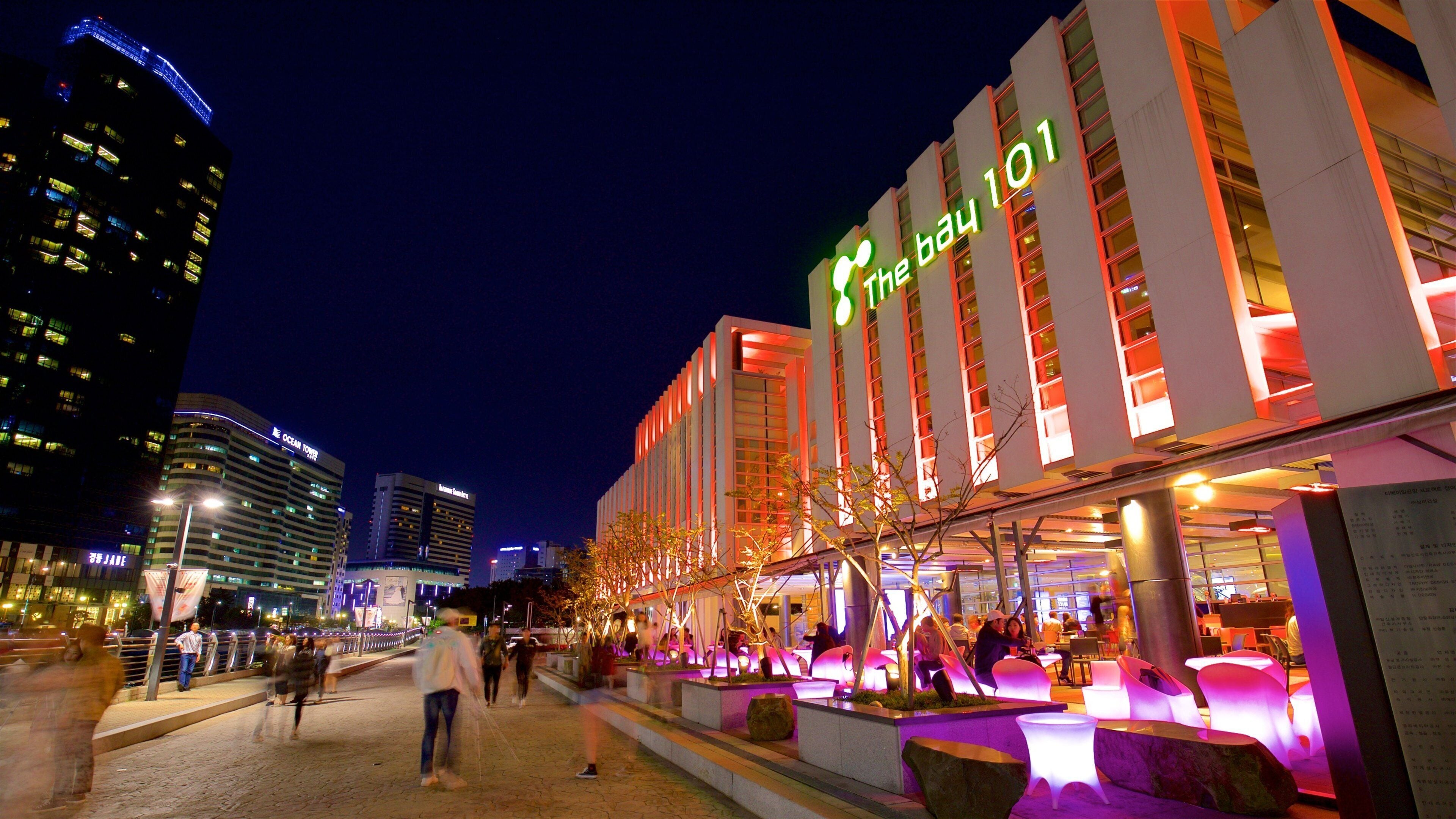 A brightly lit building with neon signage, a paved walkway, and surrounding urban buildings.