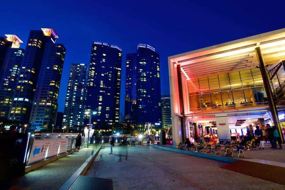 A cityscape at night with illuminated high-rise buildings and a modern building with a glass facade.