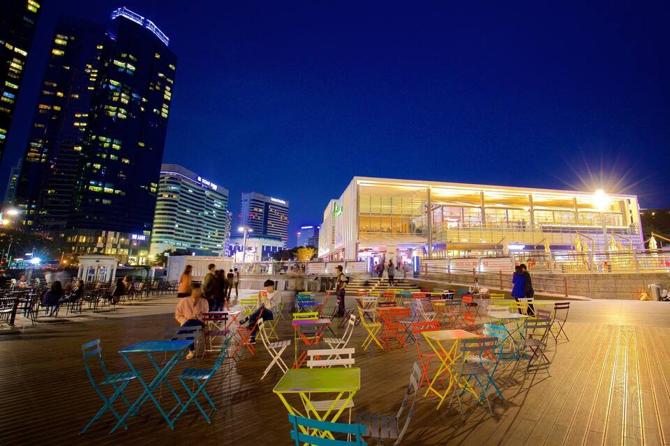 Un edificio moderno y bien iluminado con mesas de exterior coloridas en una plaza urbana de noche.