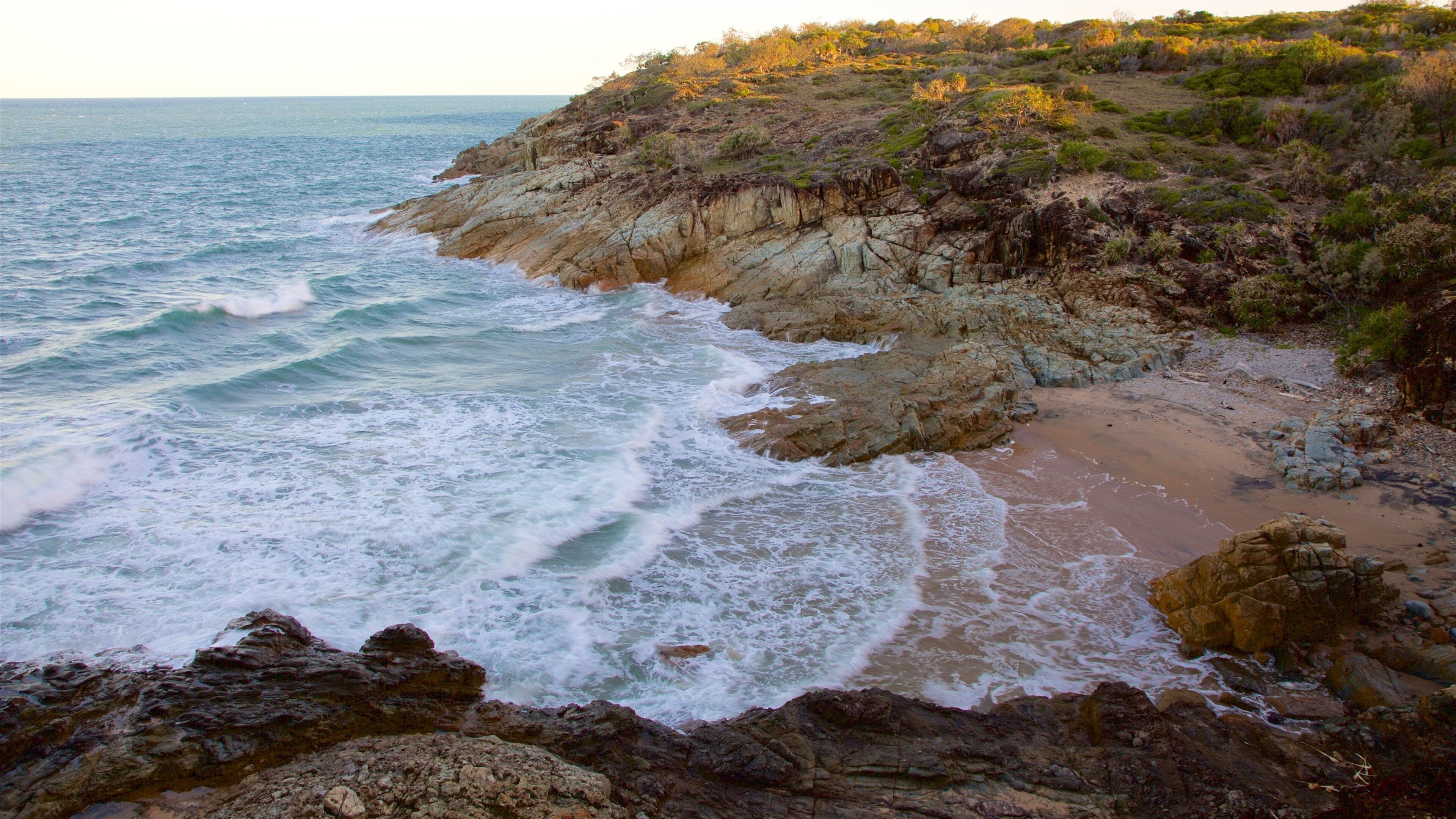 A rocky coastline with waves crashing onto the shore and a hill with sparse vegetation in the background.