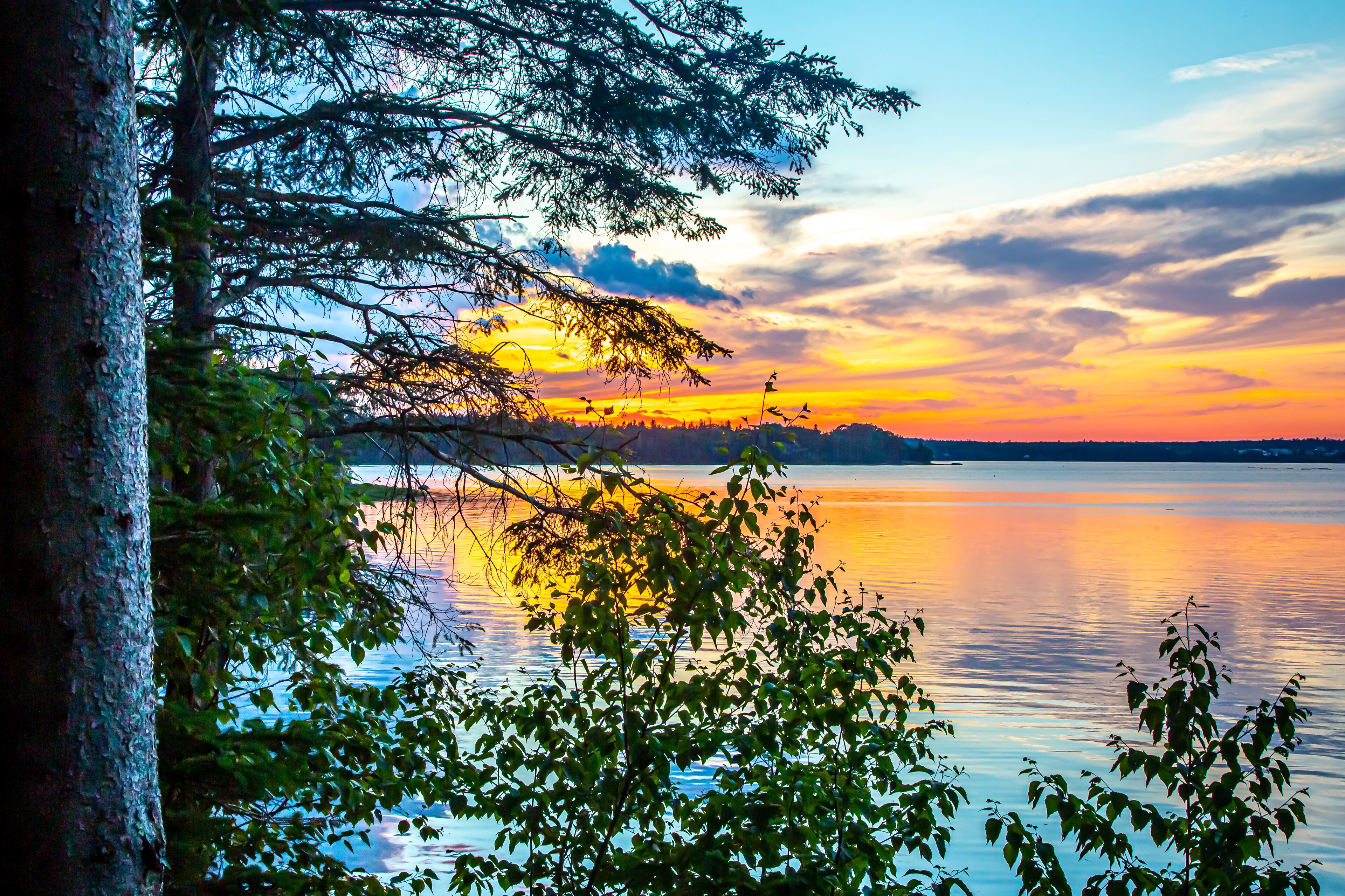 A sunset over a calm lake with trees reflecting the colors of the sky.