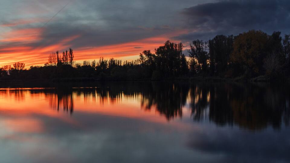A sunset over a calm lake with trees reflecting the sky.