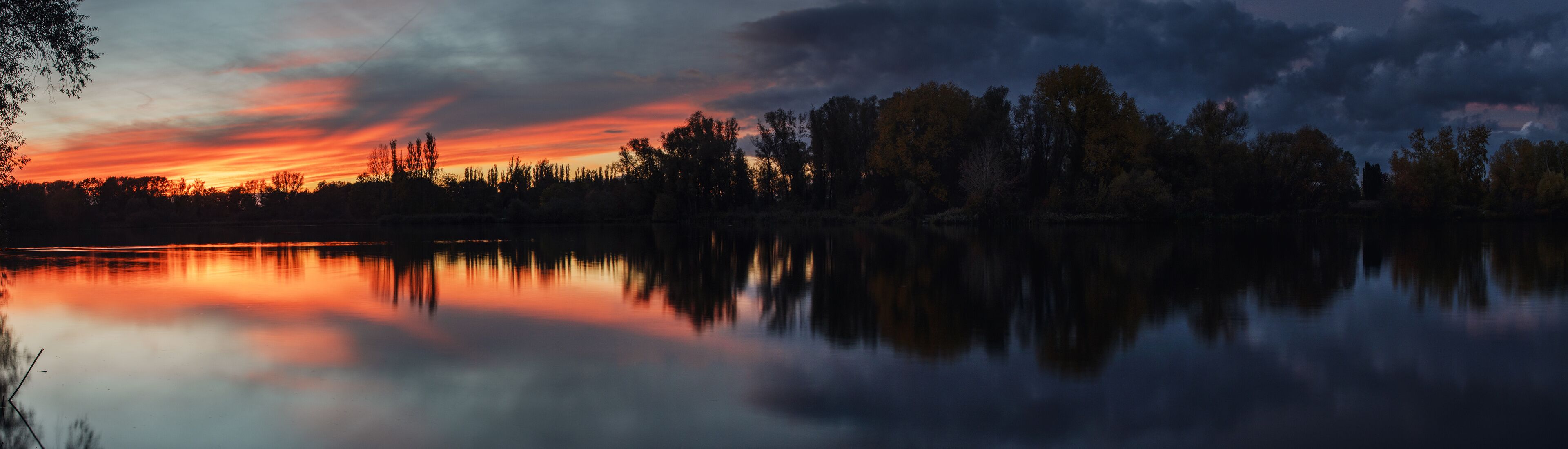 A sunset over a calm lake with trees reflecting the sky.