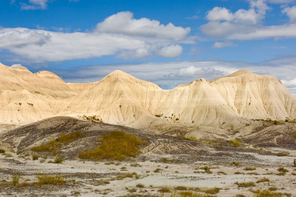 A desert landscape with rolling sand dunes and pale-colored rock formations under a partly cloudy sky.
