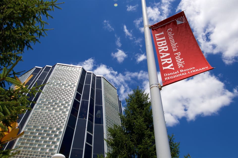 Ein modernes Gebäude mit Glasfassade und einer Flagge mit dem Aufdruck "Columbia Public Library Daniel Boone Regional Library" vor blauem Himmel mit Wolken.