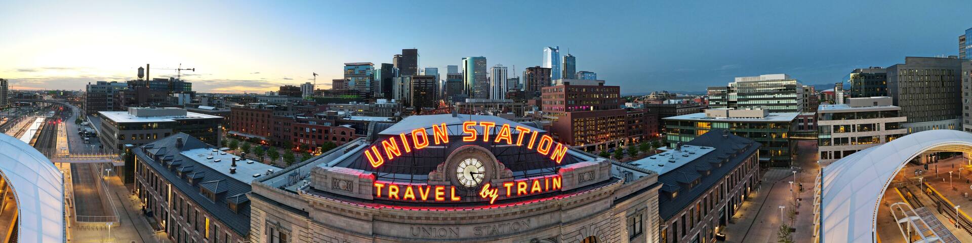union station panorama with neon travel by train sign and clock and Denver city skyline in the background at sunrise