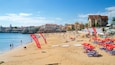 A sunny beach with red flags, people sunbathing, and a building in the background.