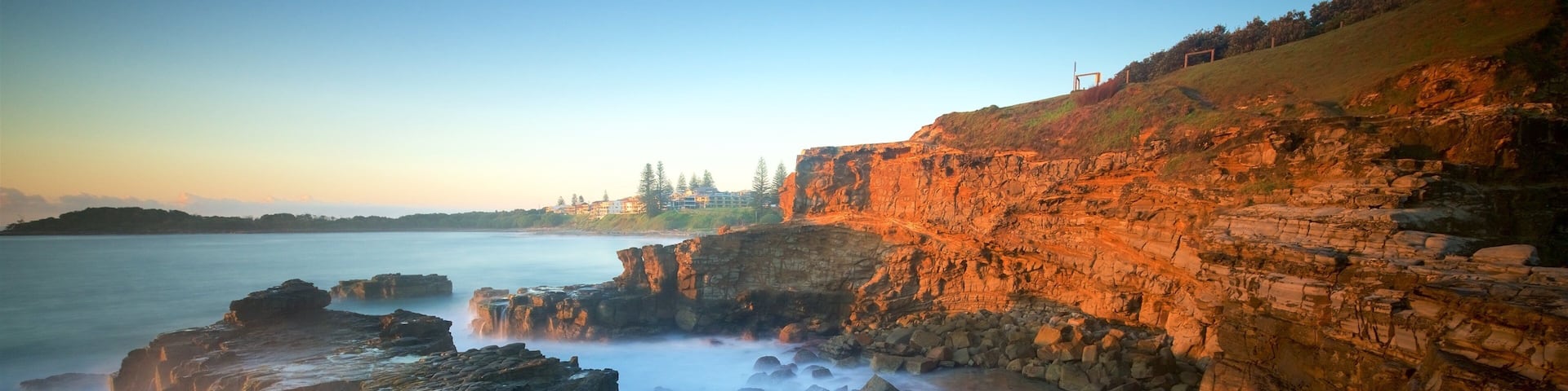 A coastal landscape with rocky cliffs, a calm sea, and a distant shoreline with buildings.