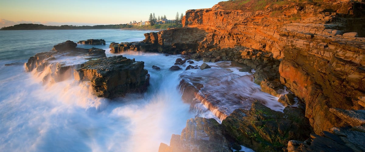 A coastal landscape with rocky cliffs, waves crashing against the rocks, and a distant shoreline with buildings.