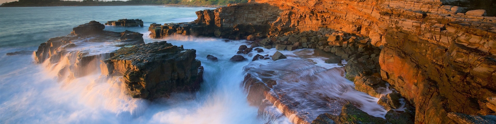 A coastal landscape with rocky cliffs, waves crashing against the rocks, and a distant shoreline with buildings.