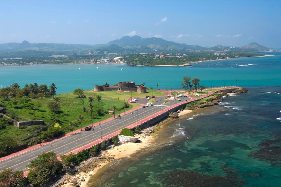 A coastal road with a historic fort, clear blue waters, and distant mountains.