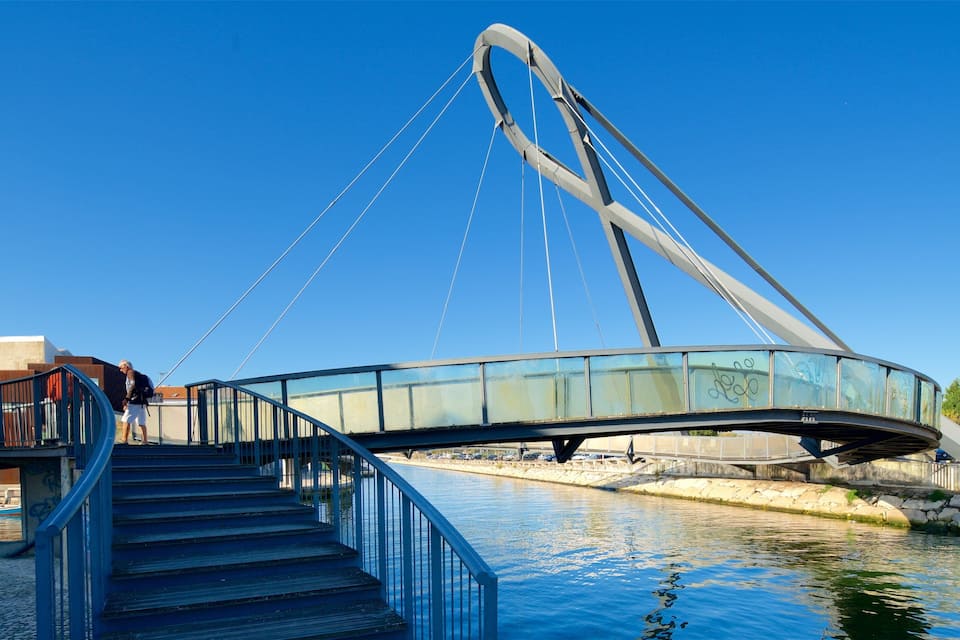 Uma moderna ponte pedonal com painéis de vidro e cabos de metal sobre um rio calmo.