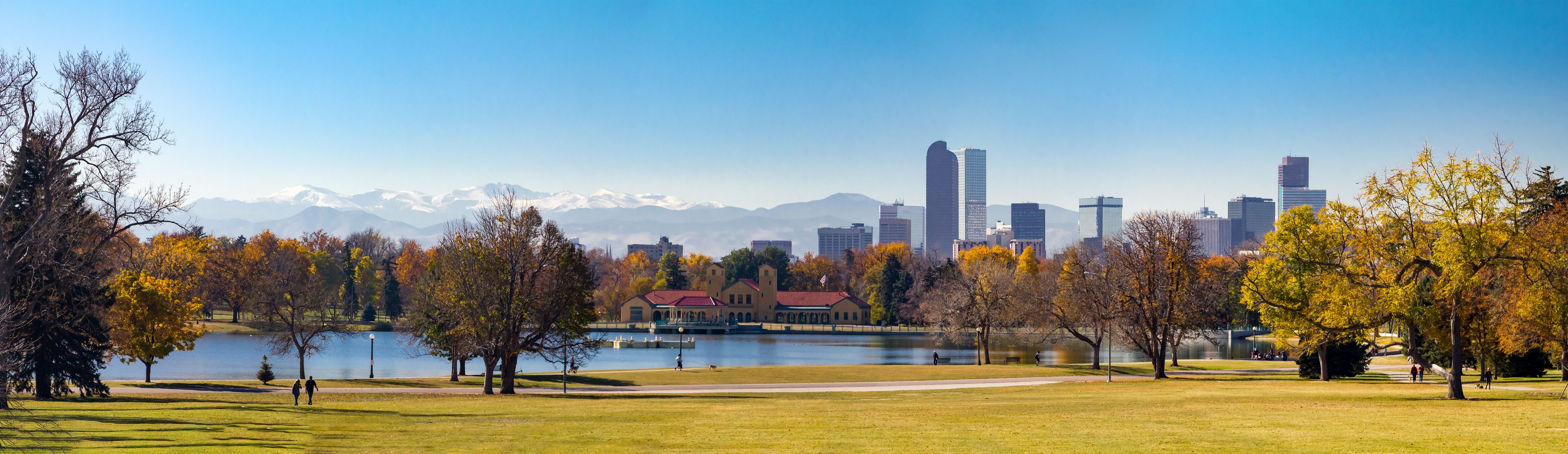 Um parque com um lago, árvores de outono e um horizonte urbano com montanhas ao fundo.