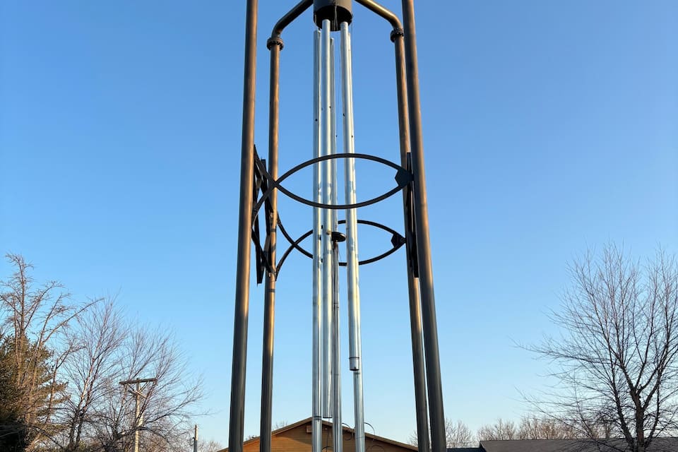 Un'alta scultura eolica in metallo in un giardino curato, con un edificio recante la scritta 'WILSON'S LARGEST WIND'.