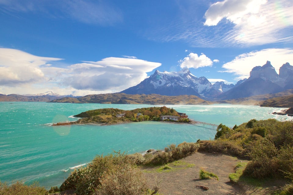 A coastal landscape with turquoise waters, a small island with houses, and snow-capped mountains in the background.