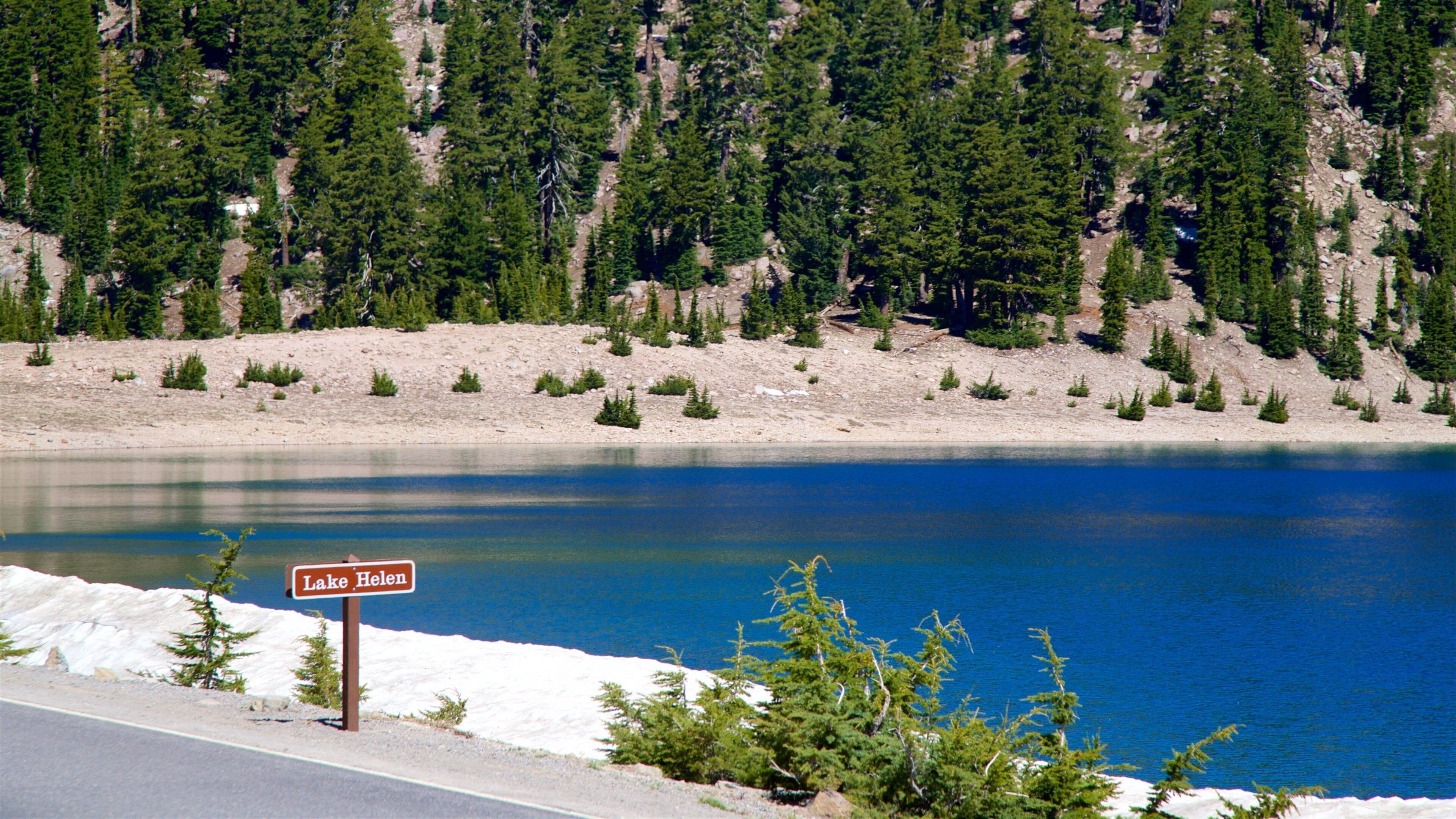 Un lac, bordé d’une pancarte indiquant « Lac Helen », entouré d’une forêt dense et d’un terrain rocheux.