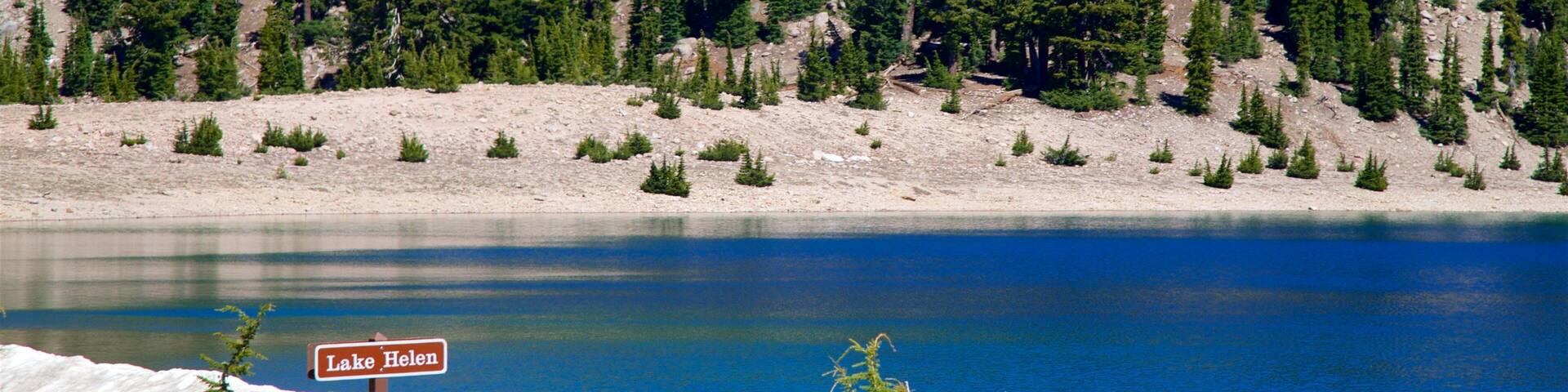 Un lago con un letrero que dice 'Lake Helen', rodeado de un bosque denso y terreno rocoso.