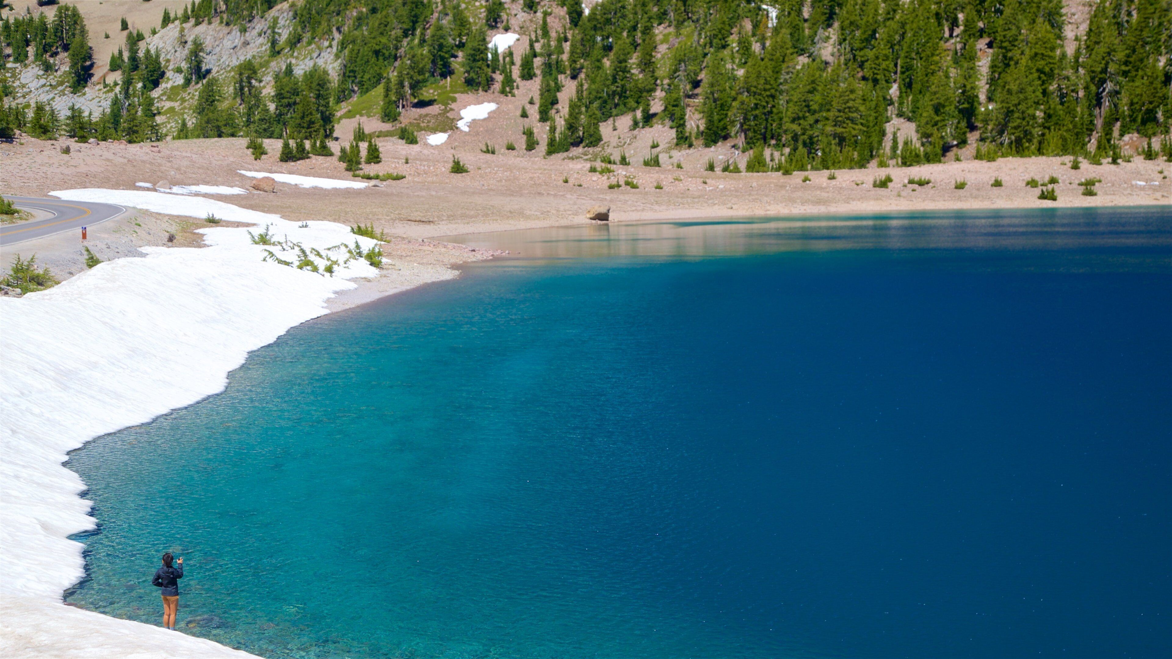 A clear blue lake with snow-capped shores and a forested mountain in the background.