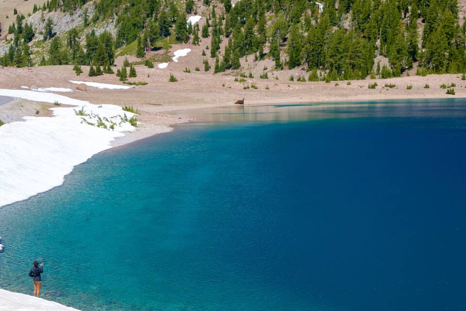 A clear blue lake with snow-capped shores and a forested mountain in the background.