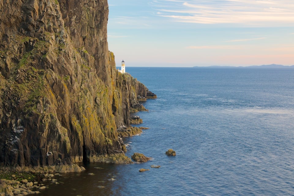 A coastal cliff with a lighthouse, rocky shoreline, and calm sea.