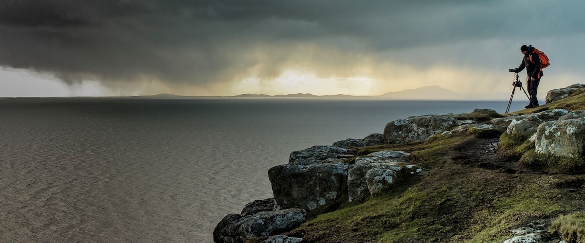 A hiker on a rocky outcrop overlooking a body of water with a stormy sky in the background.