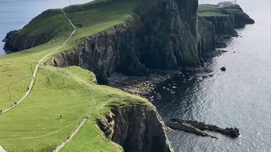 A coastal landscape with a prominent cliff, a lighthouse, and a grassy path along the cliff's edge.