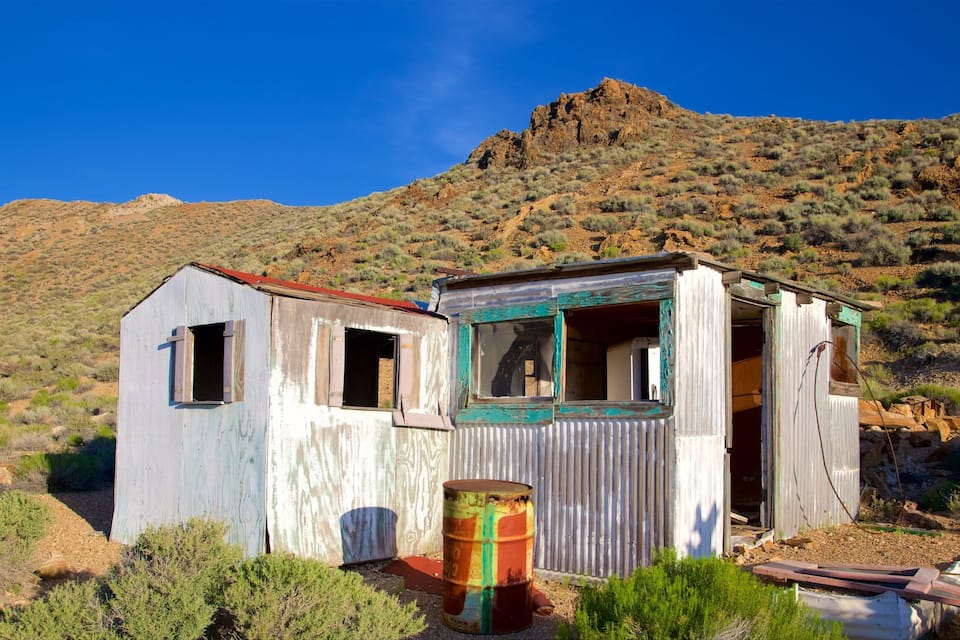 A dilapidated wooden structure with a corrugated metal roof, surrounded by sparse vegetation and a mountainous backdrop.