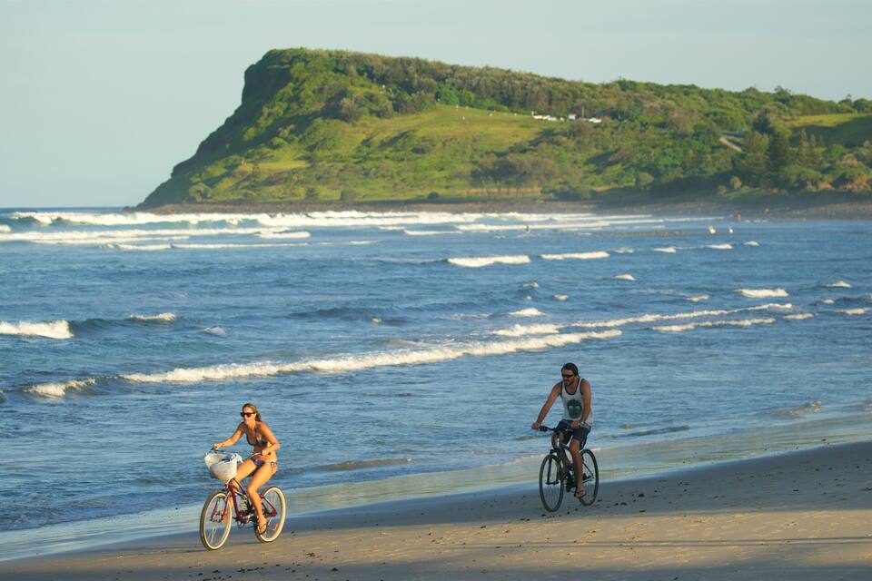 Two people riding bicycles on a beach with waves crashing onto the shore and a green hill in the background.