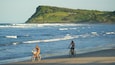 Twee personen fietsen op een strand, met golven die op het strand breken en een groene heuvel op de achtergrond.