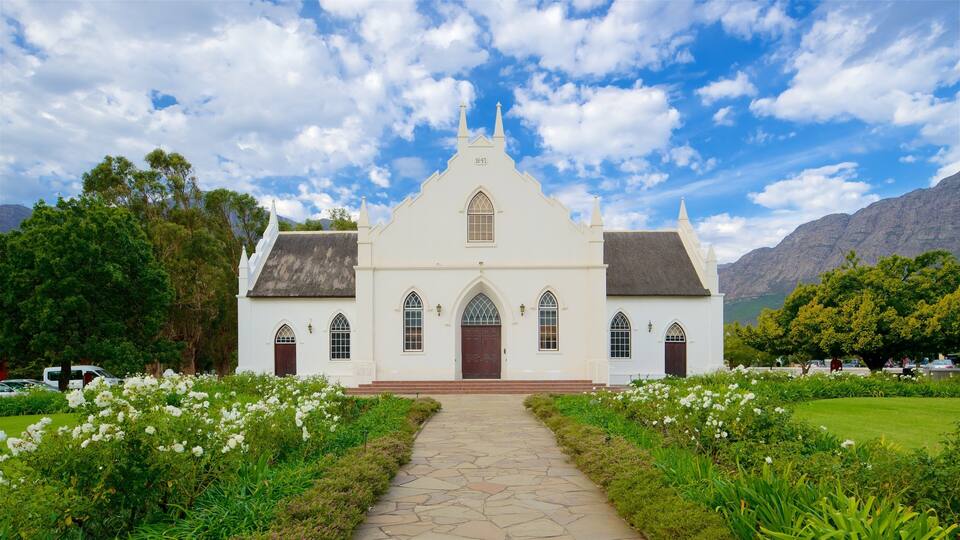 A white church with a stone pathway leading to it, surrounded by lush greenery and mountains in the background.