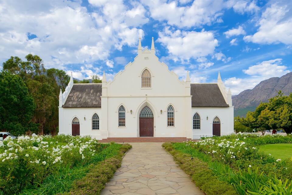 Een witte kerk met een stenen pad dat erheen leidt, omgeven door weelderige begroeiing en bergen op de achtergrond.