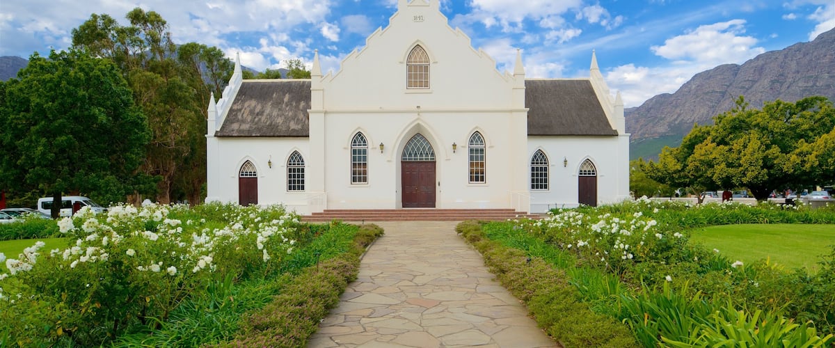 A white church with a stone pathway leading to it, surrounded by lush greenery and mountains in the background.