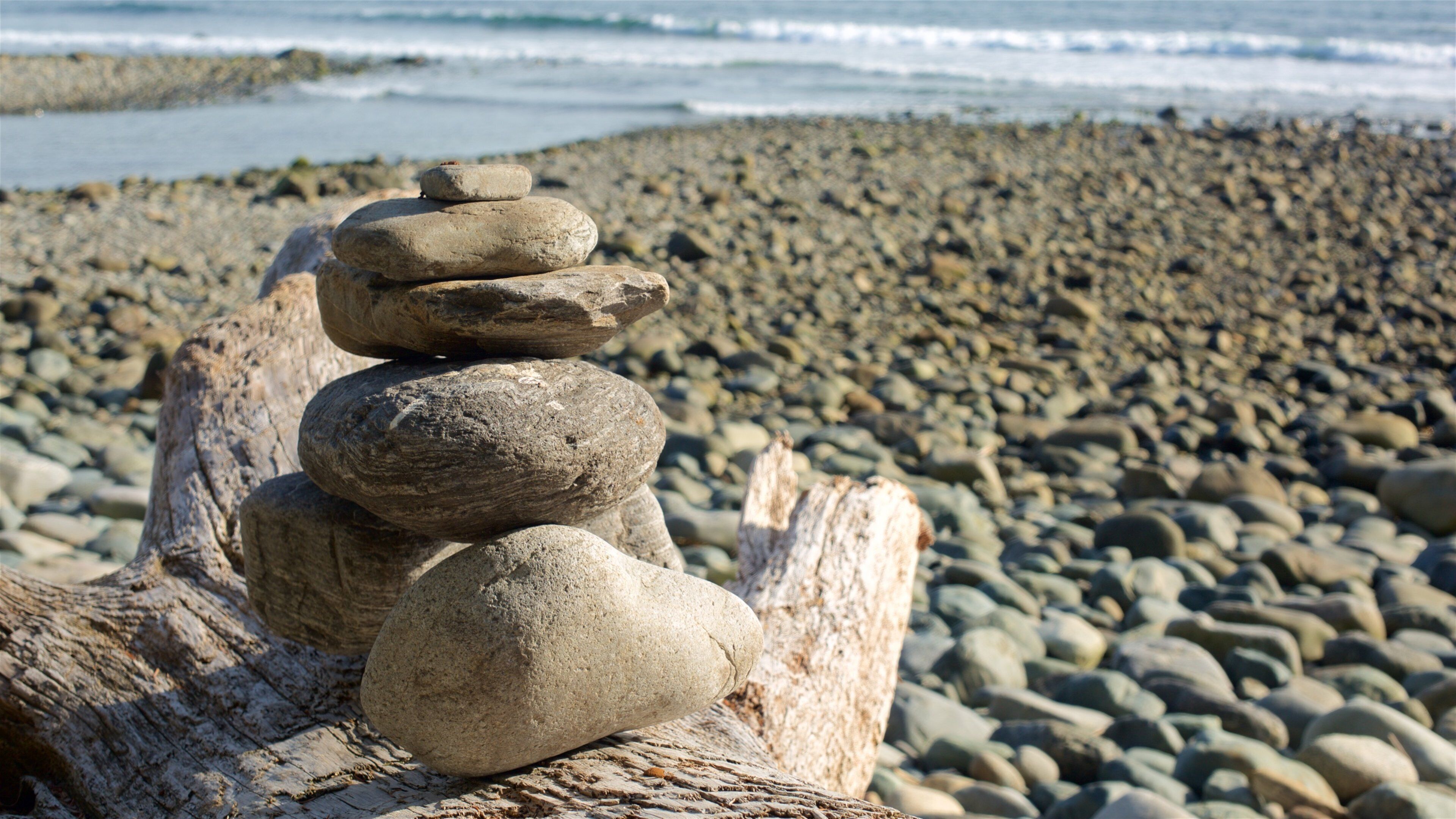 A stack of smooth, rounded stones balanced on a driftwood log by the ocean.