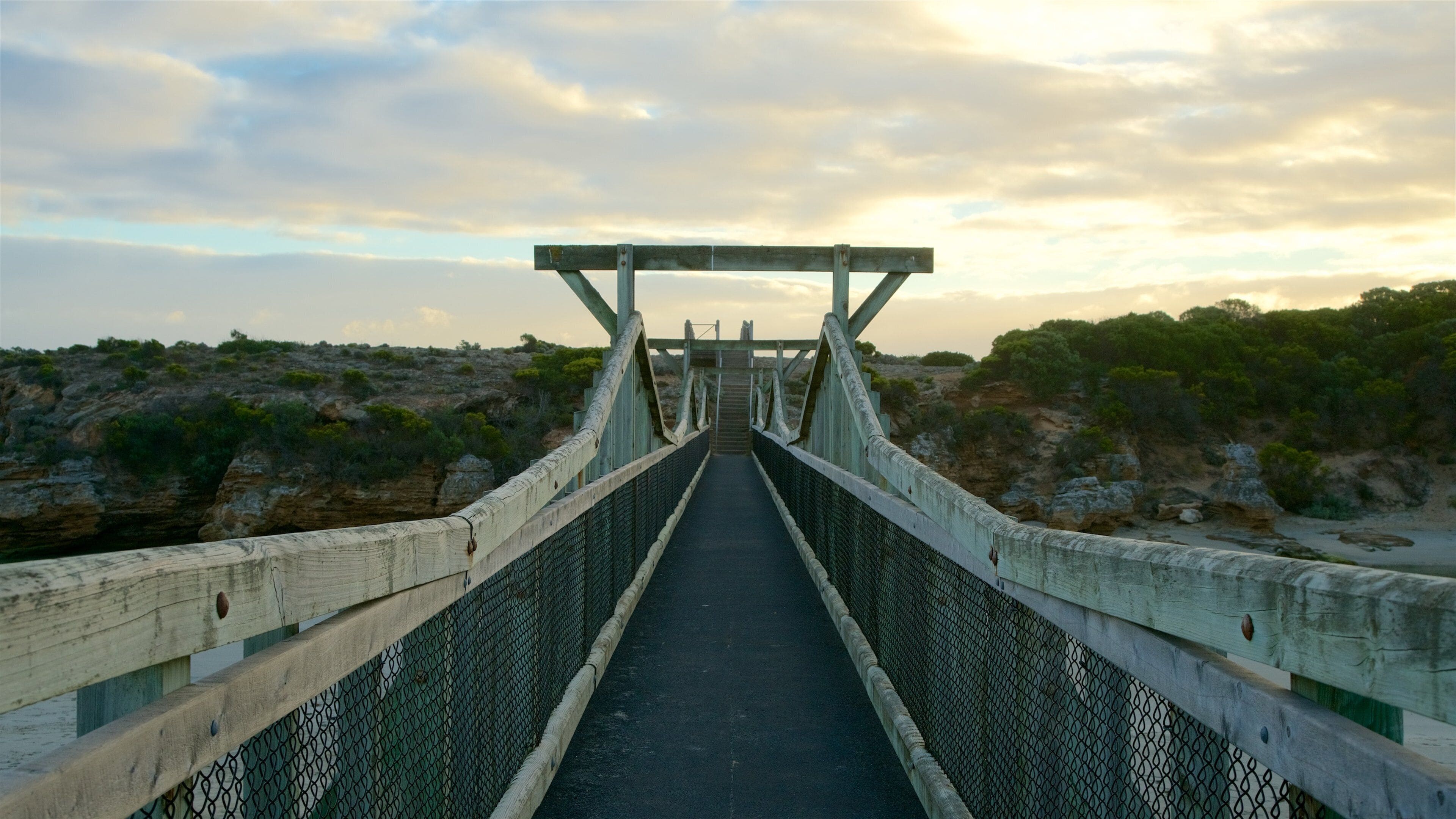 Un ponte sospeso con passerella in metallo e ringhiere in legno, che conduce verso un paesaggio roccioso e scarsamente vegetato.