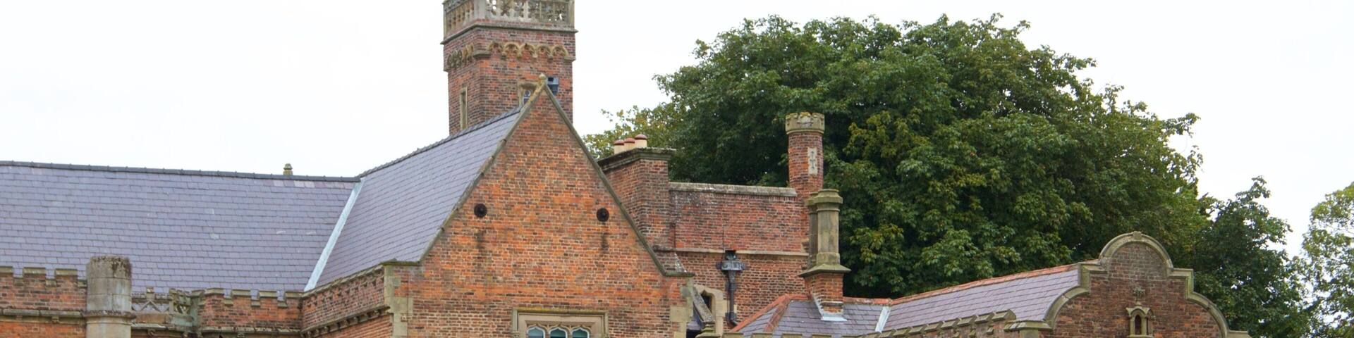 A historic brick building with a steeply pitched roof, a prominent chimney, and a large tree in the background.
