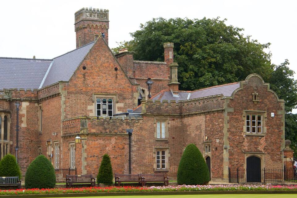 A historic brick building with a steeply pitched roof, a prominent chimney, and a large tree in the background.
