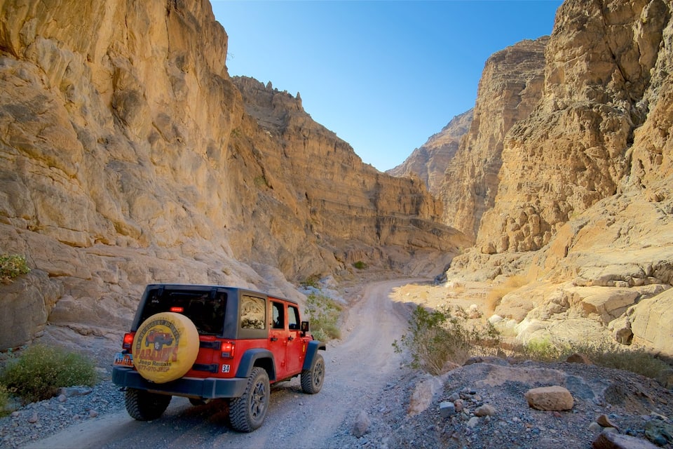 A red Jeep navigating a narrow canyon road flanked by steep, rocky cliffs.