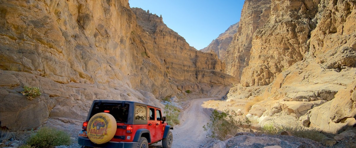 A red Jeep navigating a narrow canyon road flanked by steep, rocky cliffs.