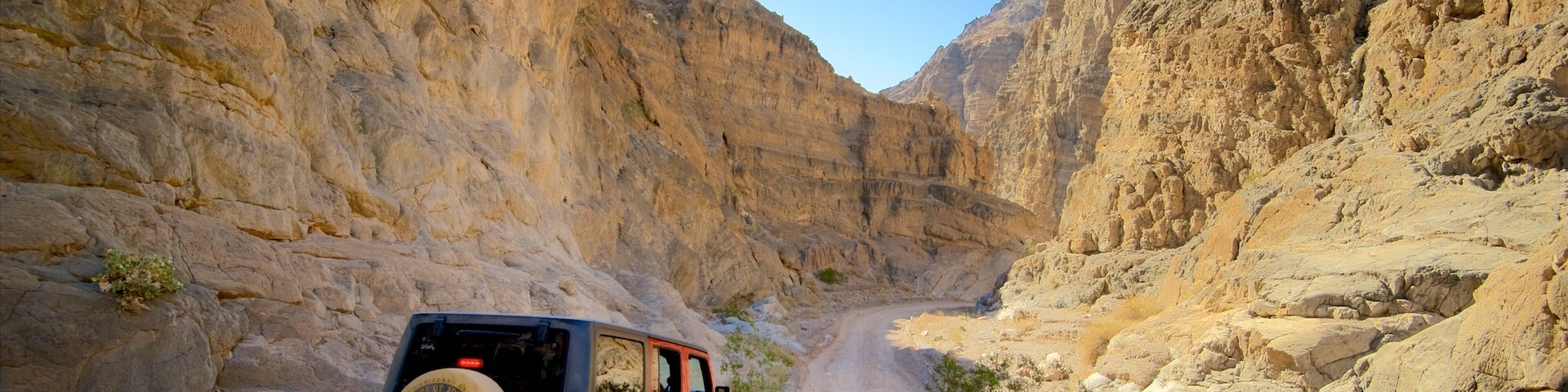 A red Jeep navigating a narrow canyon road flanked by steep, rocky cliffs.