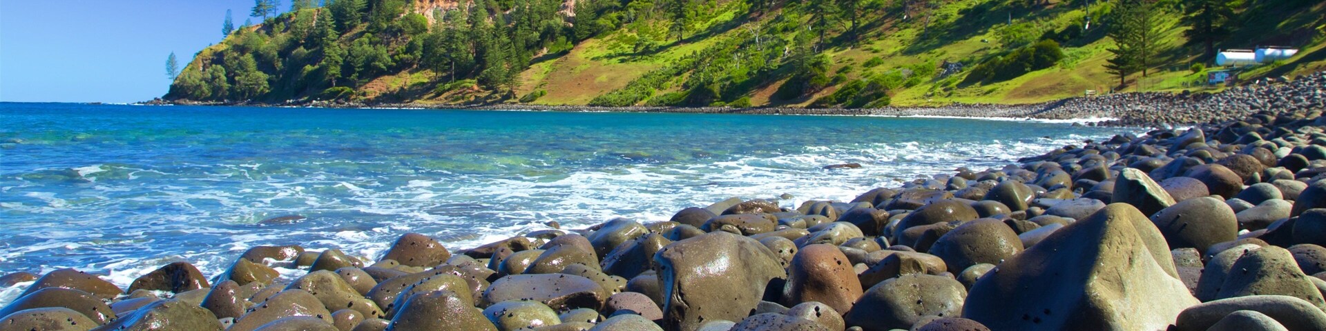 Uma costa rochosa com grandes pedras, um oceano azul límpido e um morro arborizado com edifícios ao fundo.
