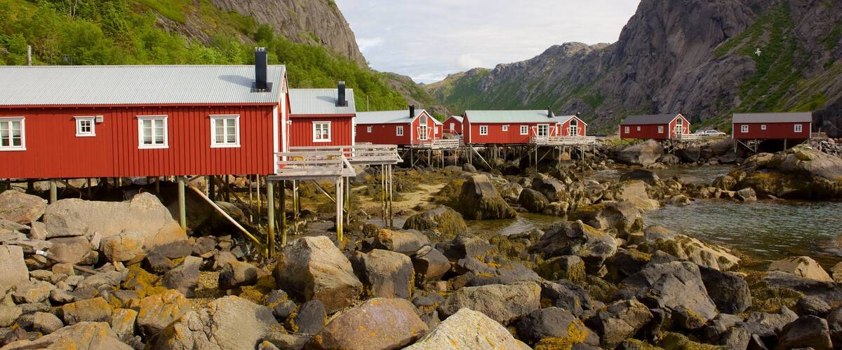 A coastal village with red wooden houses on stilts, a rocky shoreline, and mountains in the background.
