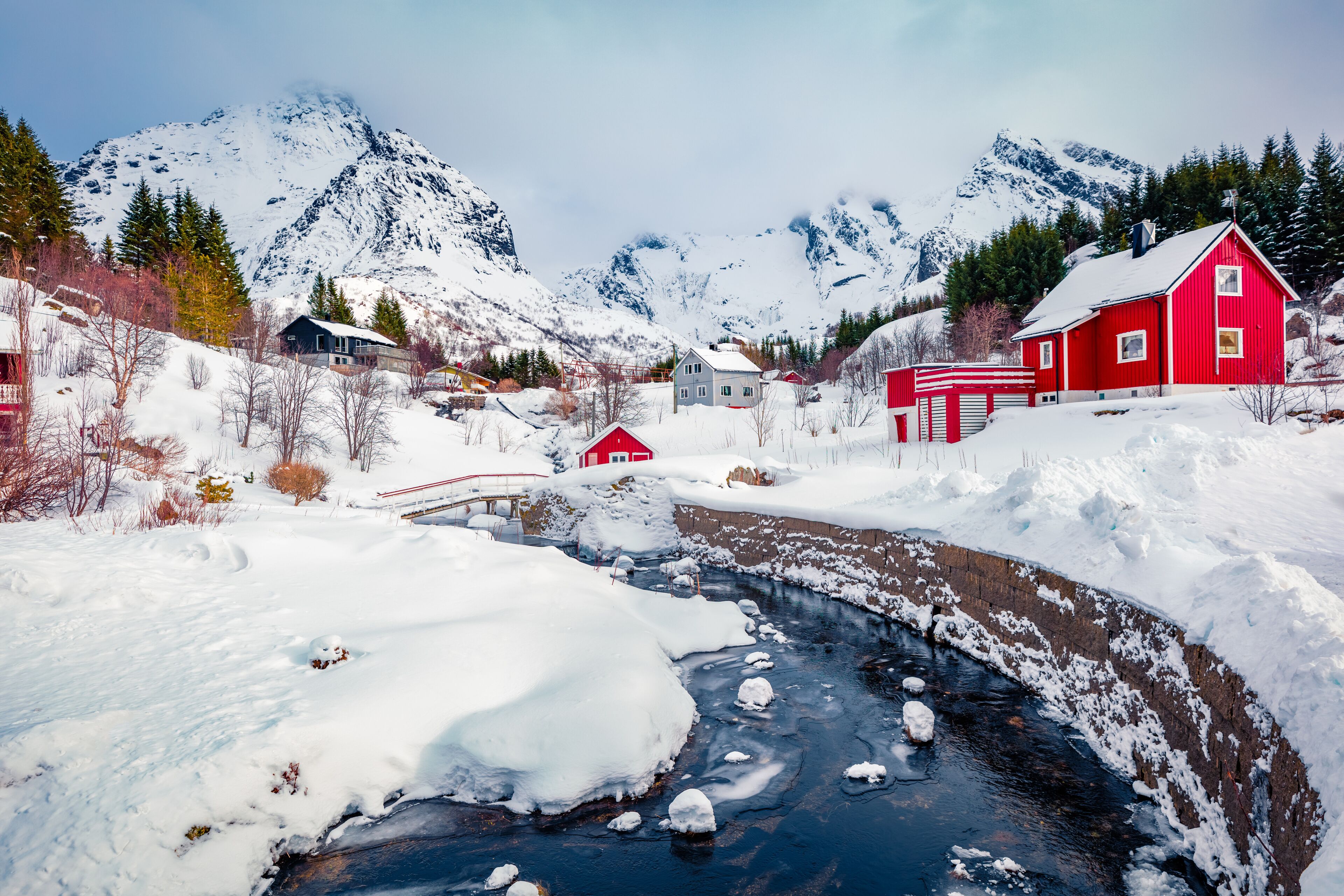 Un paisaje nevado con un río, casas rojas y montañas al fondo.