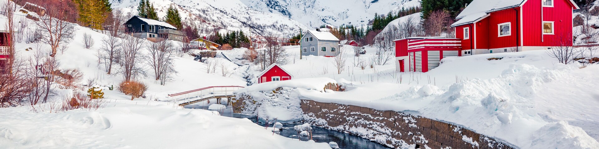 Un paisaje nevado con un río, casas rojas y montañas al fondo.
