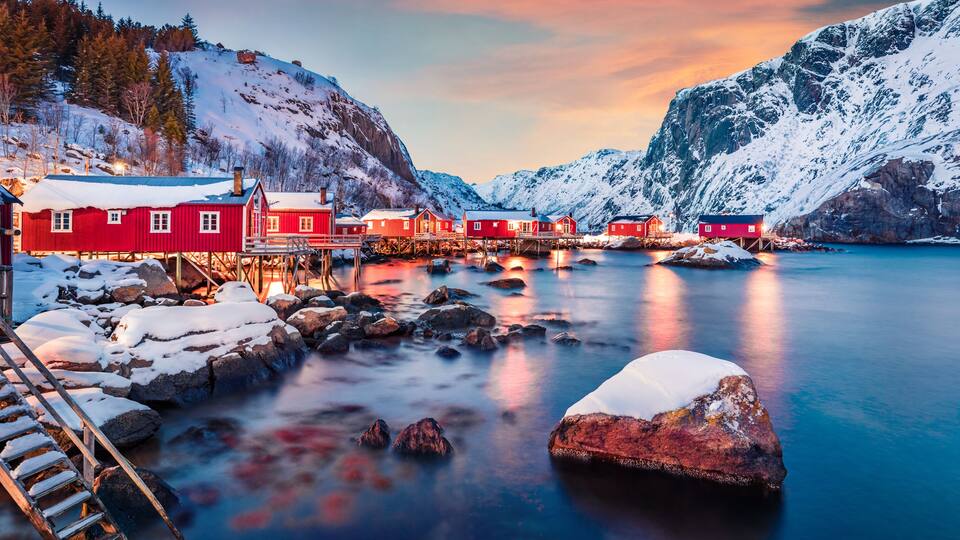 A fjord with red houses on stilts, snow-covered mountains, and a calm water surface.