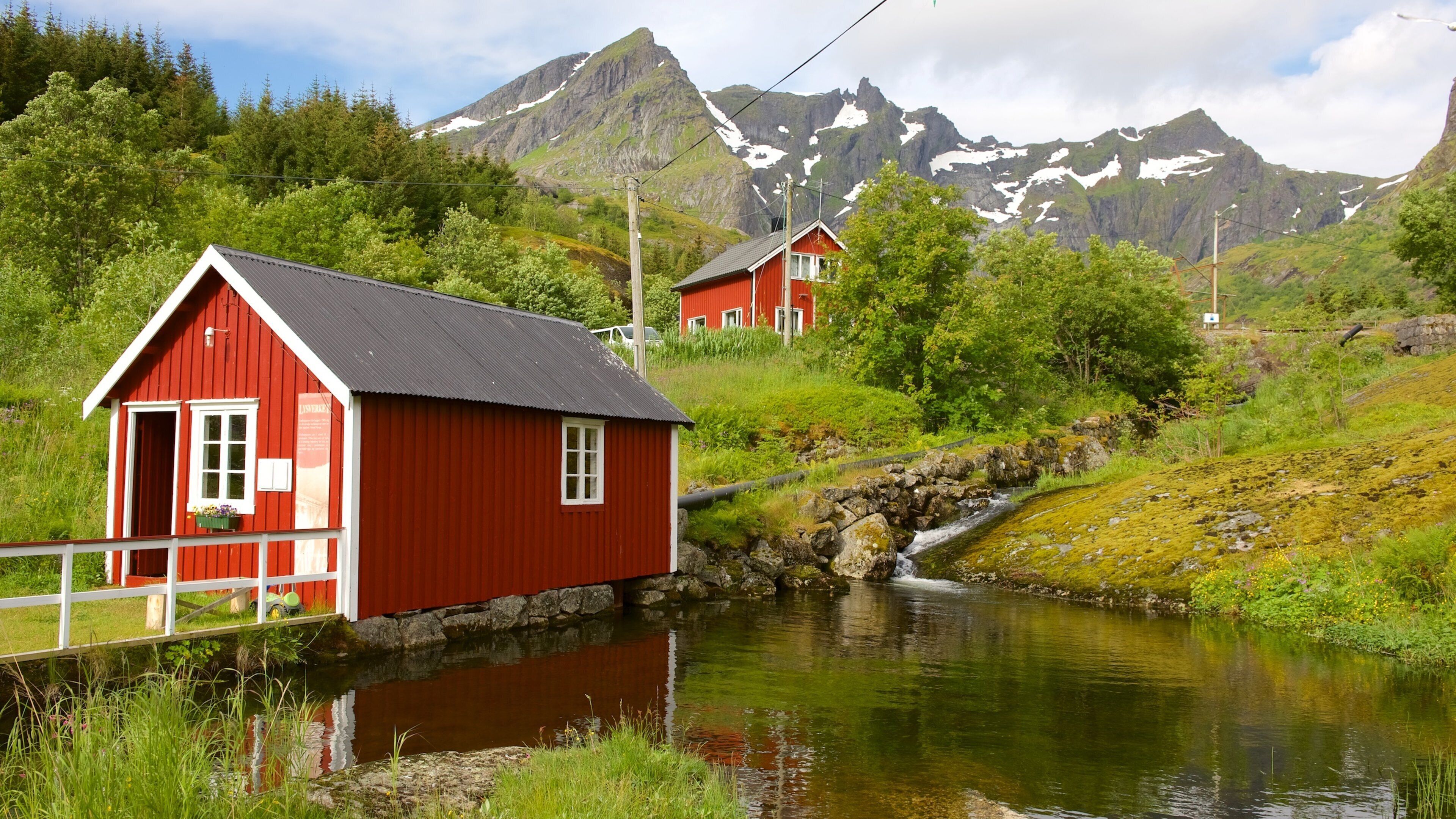A red wooden house by a calm stream with mountains in the background.