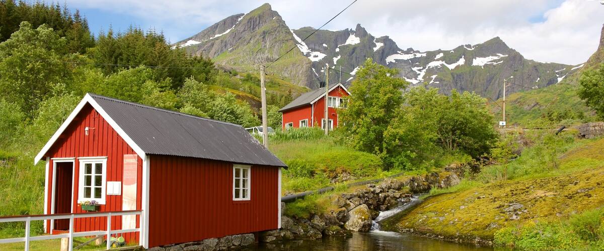 A red wooden house by a calm stream with mountains in the background.