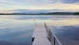 A wooden pier extending into calm water with a clear sky and distant hills.