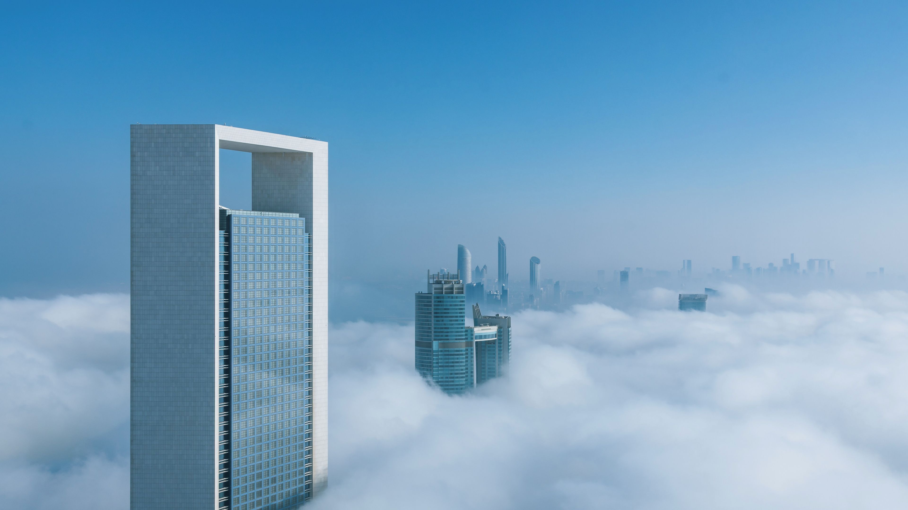 A cityscape with modern skyscrapers, one with a distinctive white and blue design, and another with a blue grid pattern, partially obscured by clouds.
